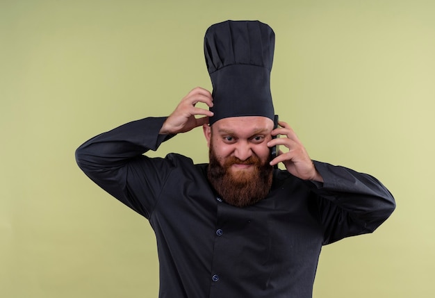 Бесплатное фото Злой бородатый шеф-повар в черной форме разговаривает по мобильному телефону с рукой за голову на зеленой стене