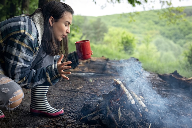 Привлекательная девушка с чашкой в руке греется у костра в лесу.