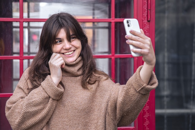 Привлекательная молодая женщина делает селфи на фоне красной телефонной будки.
