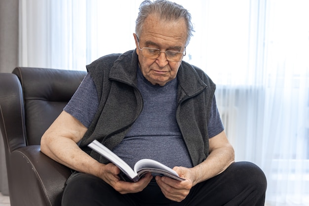 眼鏡​を​かけた​老人​が​肘掛け​椅子​に​座って​本​を​読んでいます​。