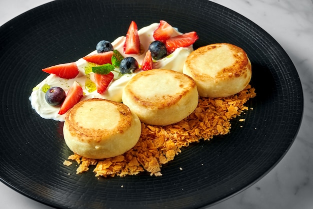 Идеальный завтрак - творожные оладьи с белым соусом, ягодами, клубникой и хрустом в черной тарелке на мраморной поверхности.