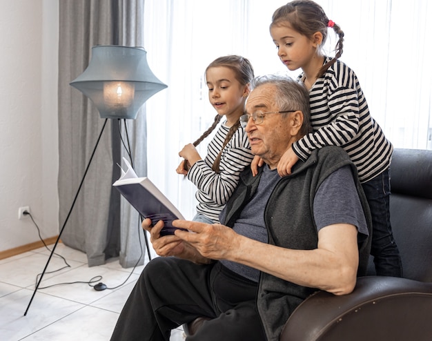 안경을 든 늙은 할아버지가 집 의자에 앉아 손녀들에게 책을 읽고 있습니다.