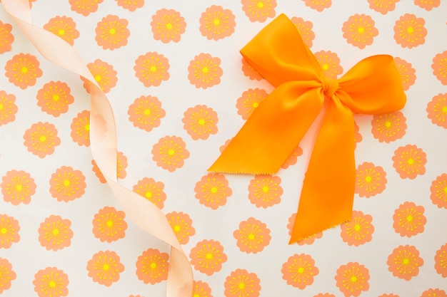 Бесплатное фото Оранжевый бант и скрученная лента на цветочной подарочной бумаге
