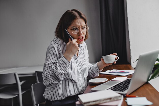 Angered businessman talking on phone with subordinates. Dissatisfied female worker in white blouse holding white cup at table with laptop.