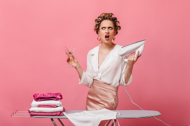 Angered woman in hair curlers holds iron and martini glass on pink wall