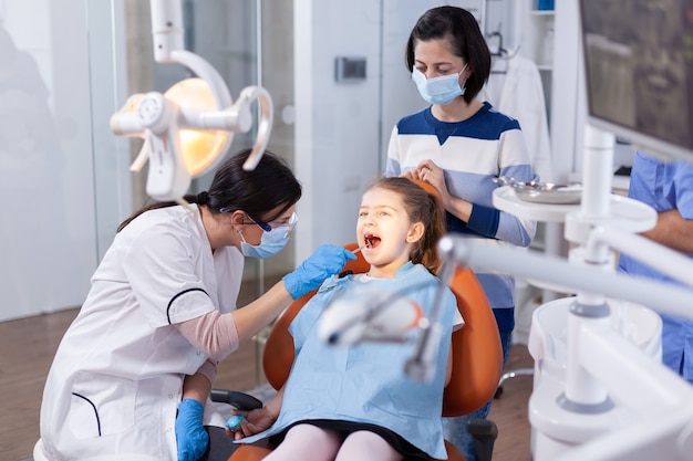 Angled mirror used by dentist doctor on little girl with mouth open in dental office. Dentistry specialist during child cavity consultation in stomatology office using modern technology.