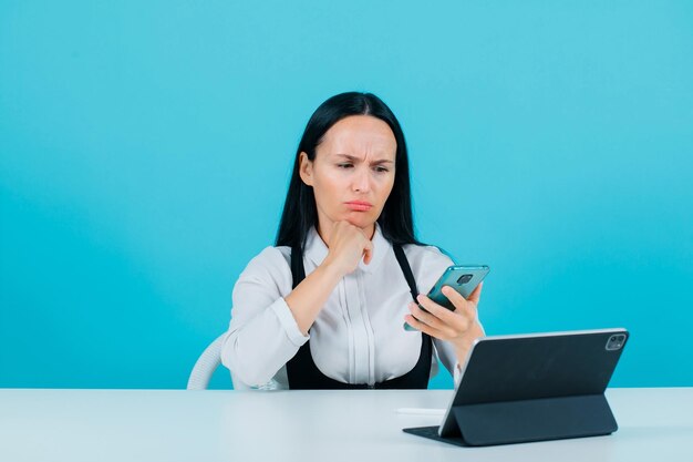 Angru blogger girl is looking at her mobile phone by sitting in front of tablet camera on blue background