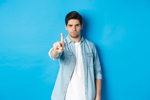 Angry 25 years old man shaking finger in disapproval sign, frowning disappointed, forbid something bad, telling no, standing against blue background