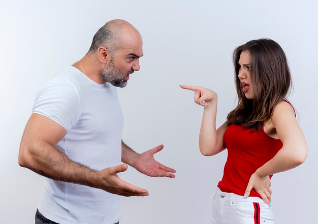 Angry adult couple arguing with each other man spreading hands and woman keeping hand on waist and pointing at him isolated on white wall