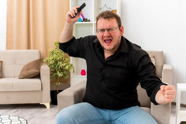 Free photo angry adult slavic man in optical glasses sits on armchair keeping fist and raising hand holding tv remote inside living room