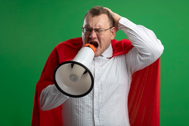 Foto gratuita arrabbiato adulto supereroe slavo uomo in mantello rosso con gli occhiali tenendo la mano sulla testa guardando il lato che grida in altoparlante isolato sulla parete verde