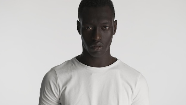 Злой афроамериканец в белой футболке выглядит агрессивно в камеру на белом фоне
