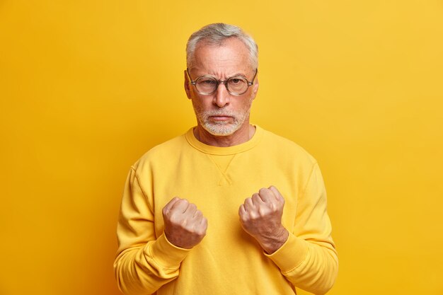 Angry aged man clenches fists as going to defense himself expresses rage and aggression looks with outraged expression at front dressed casually poses against yellow wall