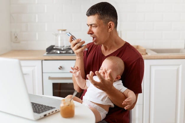 Злой агрессивный мужчина брюнетка в темно-бордовой футболке в повседневном стиле, отправляет голосовое сообщение, кричит по телефону, сидит за столом на кухне со своей младенческой дочерью.