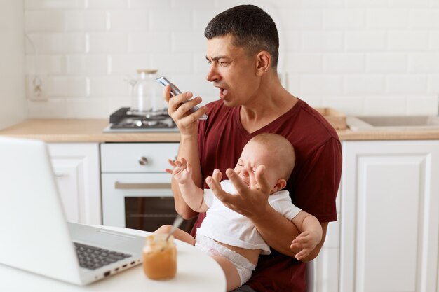 Angry aggressive brunette male wearing maroon casual style t shirt, sending voice message, screaming on telephone, sitting at table in kitchen with his infant daughter.