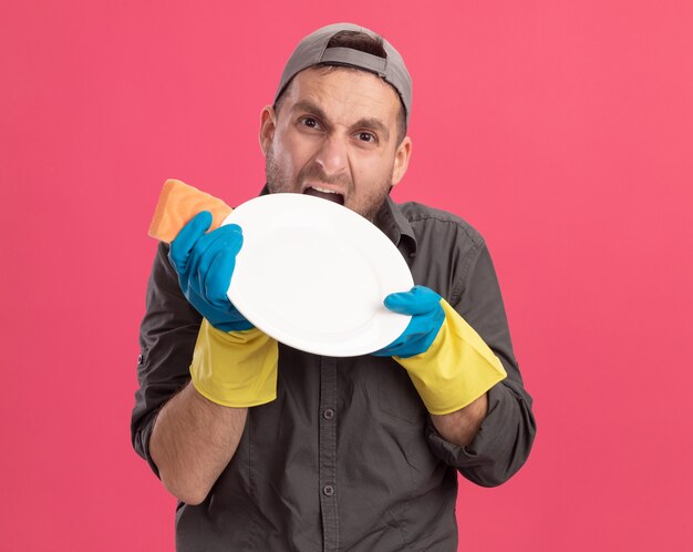 Angry and annoyed young cleaning man wearing casual clothes and cap in rubber gloves holding plate and sponge going wild biting plate standing over pink wall