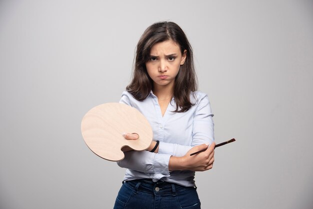 Angry artist holding paintbrush and palette over gray wall. 