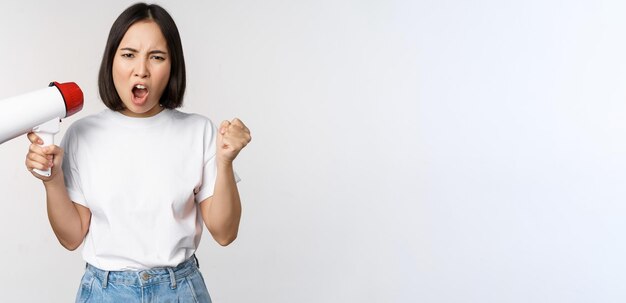 Angry asian girl activist holding megaphone and looking furious protesting standing over white background Copy space