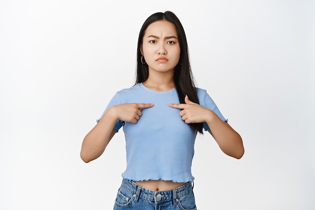 Angry asian girl pointing fingers at herself frowning and sulking upset standing in blue tshirt over white background