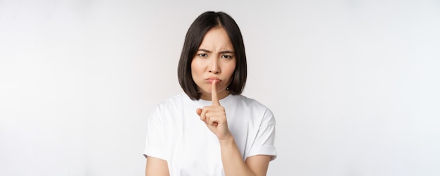 Angry asian girl shushing keep quiet taboo silence gesture press finger to lips and frowning scolding for being too loud standing over white background