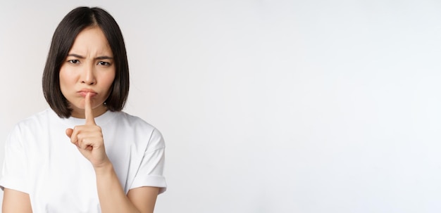 Angry asian girl shushing keep quiet taboo silence gesture press finger to lips and frowning scolding for being too loud standing over white background