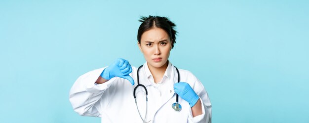 Злая азиатская женщина-врач в униформе и перчатках показывает большие пальцы вниз, нахмурив брови, расстроена дис