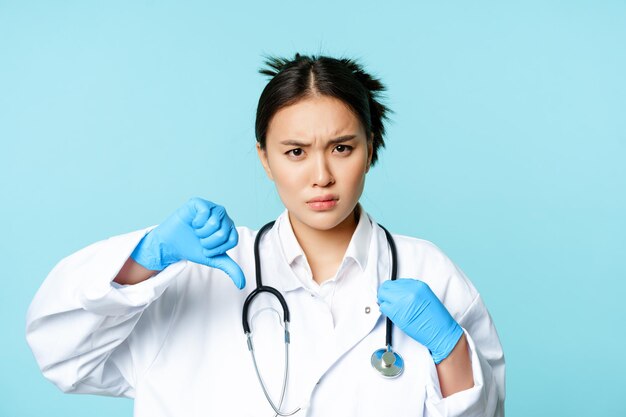 제복을 입은 화난 아시아 여성 의사 의사가 장갑을 끼고 고랑눈썹을 아래로 엄지손가락을 보여줍니다.