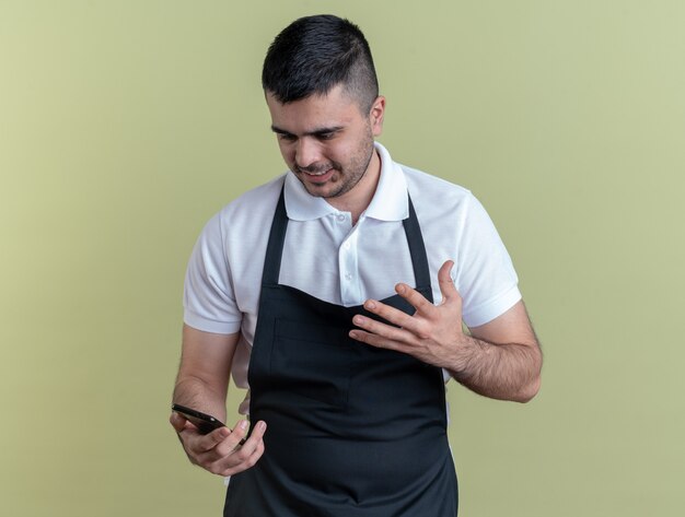 Uomo arrabbiato del barbiere in grembiule che tiene smartphone guardandolo dispiaciuto alzando il braccio indignato