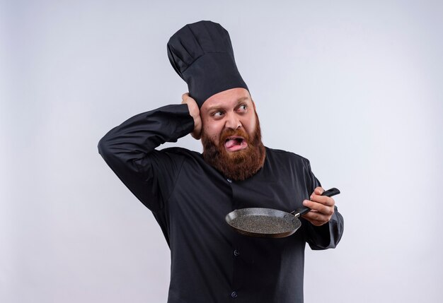 An angry bearded chef man in black uniform holding frying pan while looking side with hand on head on a white wall