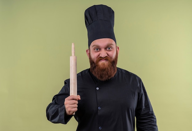 An angry bearded chef man in black uniform holding rolling pin while looking on a green wall