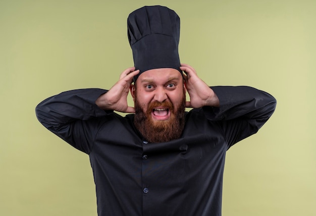 Free photo an angry bearded chef man in black uniform screaming with hands on head while looking on a green wall