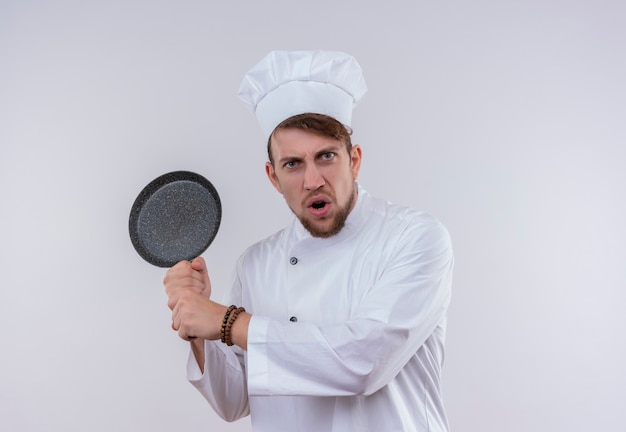 An angry bearded chef man wearing white cooker uniform and hat holding frying pan like a baseball bat on a white wall