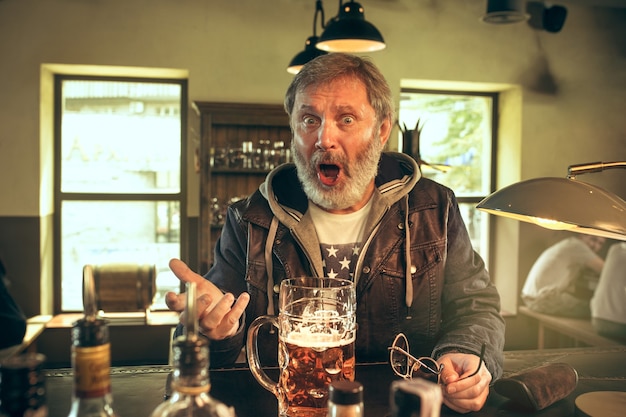 Uomo barbuto arrabbiato che beve alcolici nel pub e guarda un programma sportivo in tv. gustando il mio brulicare e la mia birra preferiti. uomo con boccale di birra seduto a tavola. appassionato di calcio o di sport. concetto di emozioni umane