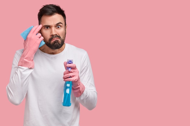 Free photo angry bearded man frowns face in discontent, has much work, cleans room, holds washing detergent in one hand and mop in other