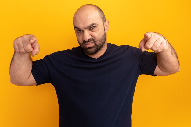 Angry bearded man in navy t-shirt pointing with index fingers  standing over orange wall