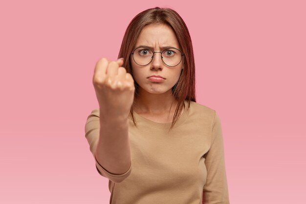 Angry beautiful young woman shows fist , threats someone, being annoyed with noisy neighbours, purses lower lip, dressed in casual clothes