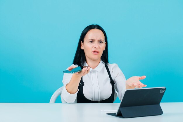 Angry blogger girl is looking at tablet camera by holding phone on blue background