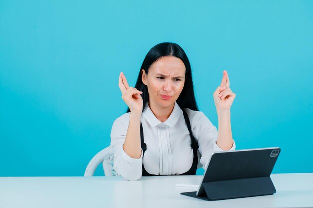 Angry blogger girl is posing to tablet camera by crossing fingers on blue background