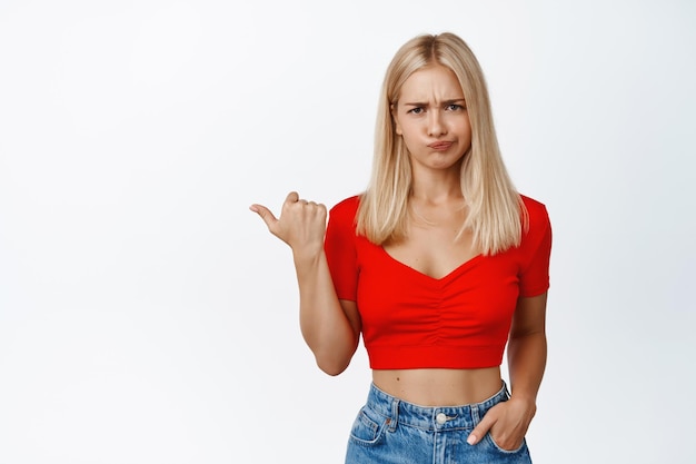 Foto gratuita ragazza bionda arrabbiata punta il dito a sinistra imbronciato e sembra scontento mostra smth bad si lamenta di pubblicità stand in top corto con jeans su sfondo bianco