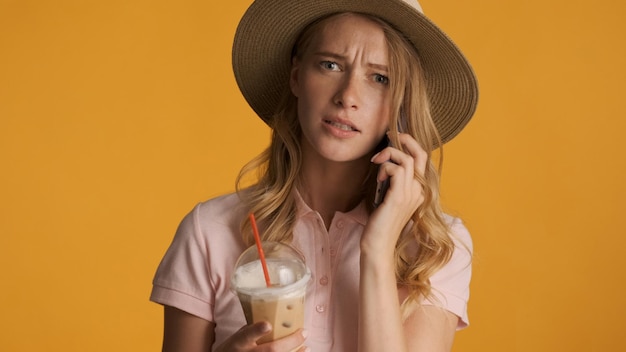 Angry blond woman in hat with coffee to go have serious phone conversation isolated on yellow background