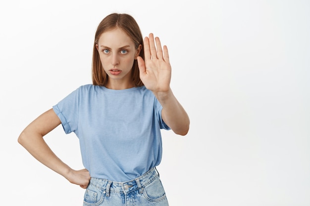 Foto gratuita donna bionda arrabbiata allunga la mano e accigliata, dice no, non è d'accordo, rifiuta o rifiuta, proibisce l'azione, non è d'accordo con la persona, in piedi sul muro bianco