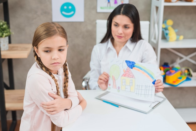 An angry blonde little girl not looking at drawing paper shown by her psychologist