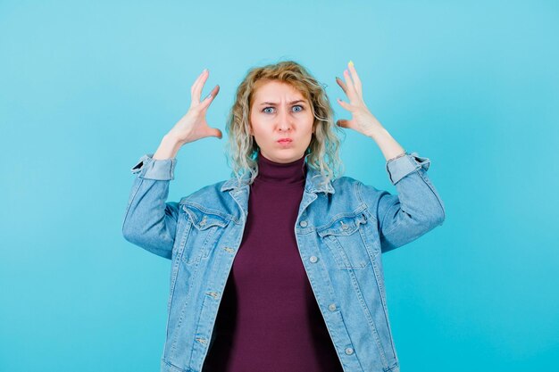 Angry blonde woman is looking at camera by holding hands near head on blue background