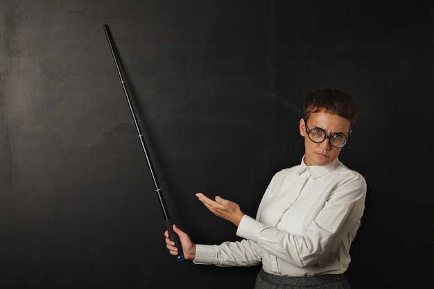 Angry brunette coach in business suit with bad emotion and shows on black chalk board behind her with folding pointer and other hand.