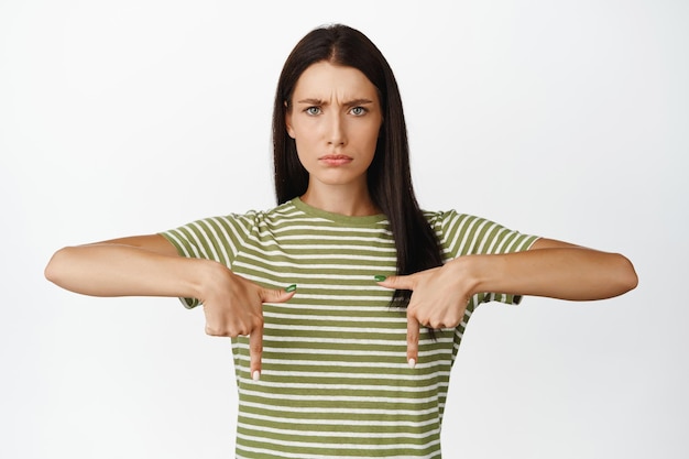 怒っているブルネットの女性は、白い背景の上のTシャツに立っているsmthで眉をひそめていると指さし指をやめます