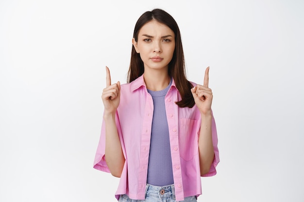 Angry brunette woman furrow eyebrows pointing fingers up dislike something standing over white background