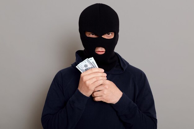 Angry burglar guy looks at the front with a forged expression and holds the stolen money in his hands