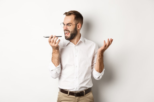 Angry business man shouting at speakerphone, record voice message in mad state, standing  