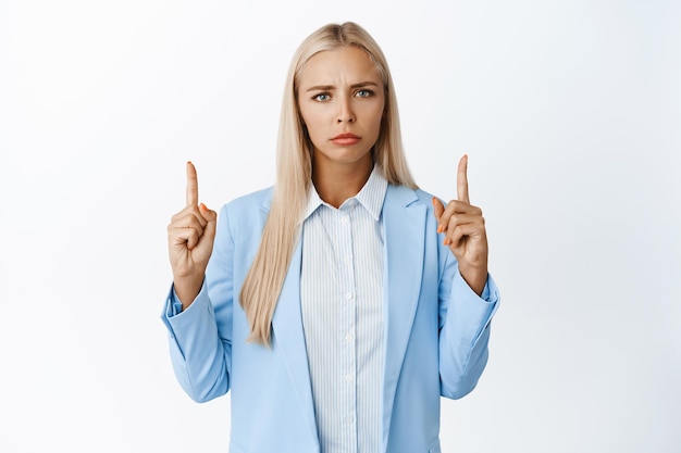 Angry businesswoman pointing fingers up frowning and looking with disappointment standing in stylish suit over white background