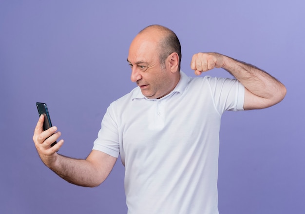 Angry casual mature businessman holding and looking at mobile phone and raising up fist isolated on purple background